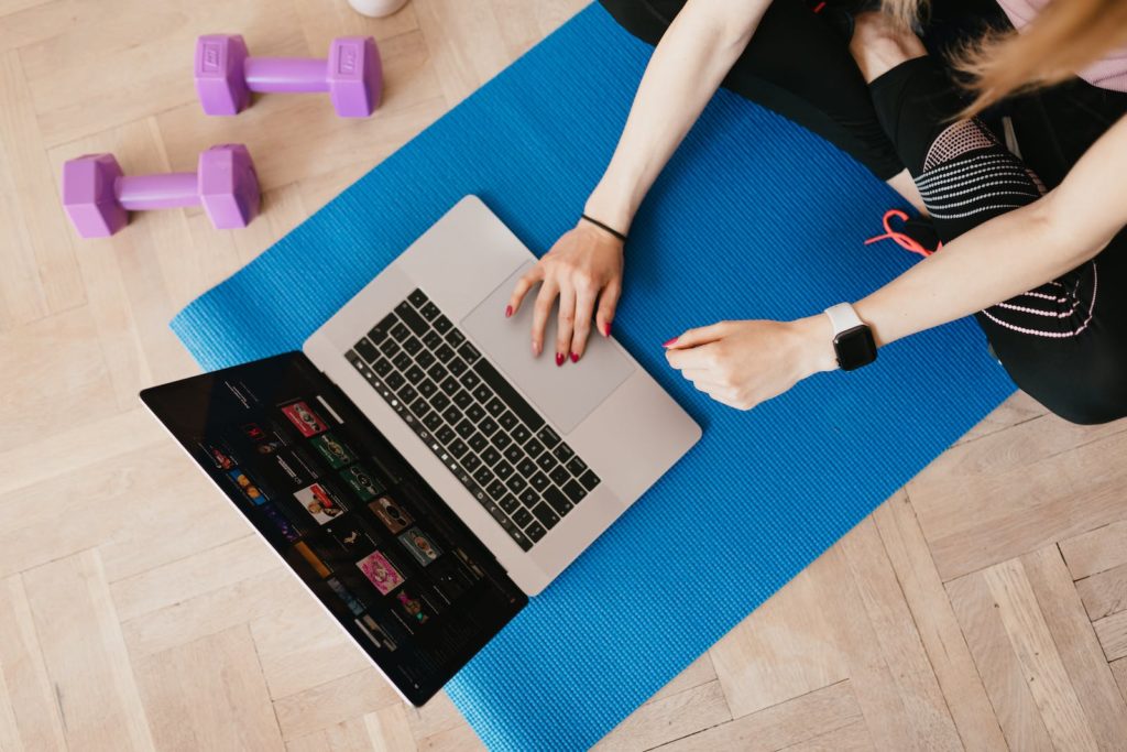 Top view of crop female in activewear sitting on yoga mat on floor and using laptop