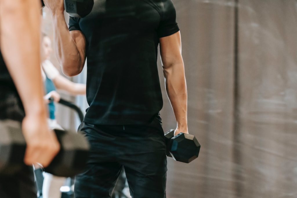 Unrecognizable muscular male in black sportswear pumping biceps with heavy metal dumbbells near mirror during workout in modern fitness studio